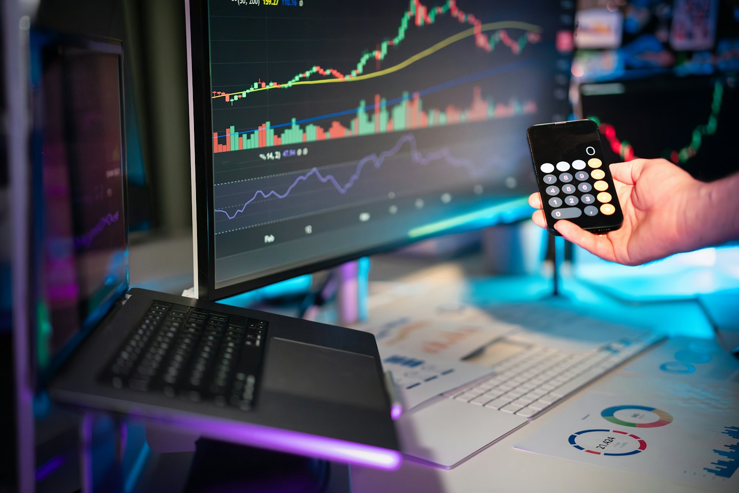 Dłoń trzyma telefon z kalkulatorem w pobliżu danych finansowych na monitorach.